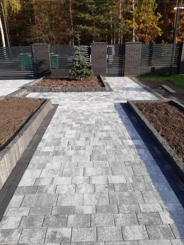 Szara kostka brukowa ułożona wzdłuż ścieżki z wyznaczonymi rabatami, w tle nowoczesne, ciemne ogrodzenie z elementami z cegły.