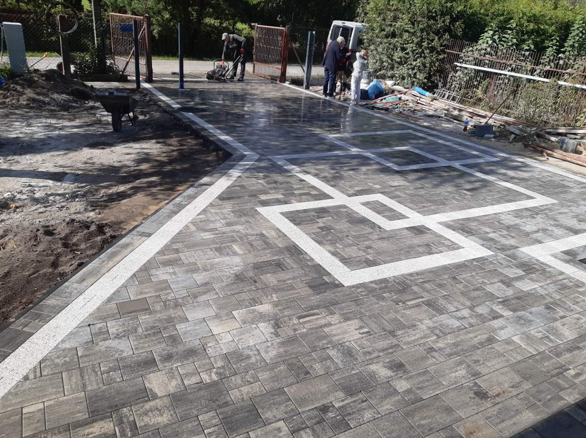 Ułożona nawierzchnia z szarej kostki brukowej z białymi, dekoracyjnymi liniami tworzącymi geometryczny wzór; w tle pracownik zagęszcza teren ubijarką, a w oddali ekipa montuje bramę wjazdową.