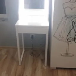 Biała toaletka z podświetlanym lustrem stoi na drewnianej podłodze, obok szafy z rysunkiem manekina.