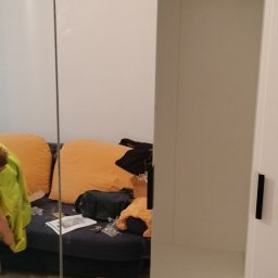 Sławek Kozik - Biała szafa z przesuwnymi lustrzanymi drzwiami, widoczne wnętrze z półkami i szufladami, odbicie pokoju z sofą i porozrzucanymi przedmiotami.