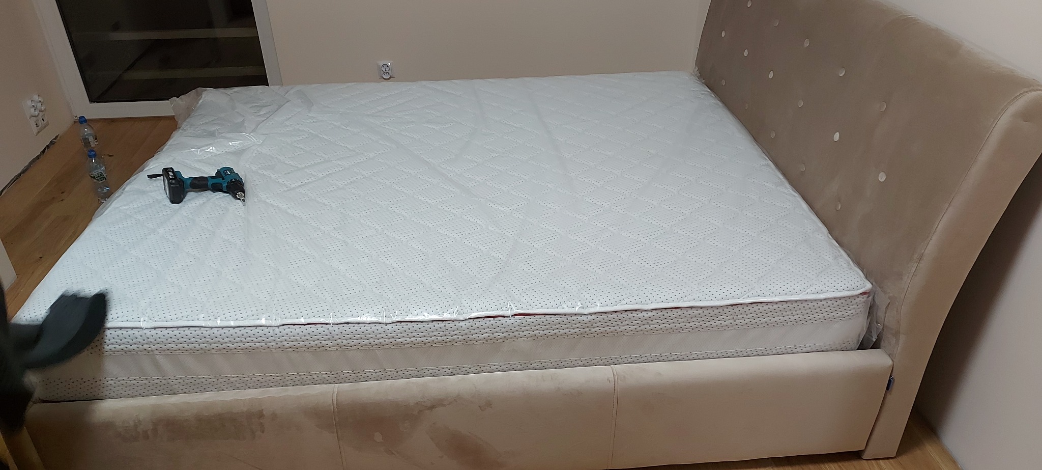 Beżowe łóżko z materacem w folii ochronnej, wiertarka akumulatorowa leży obok, butelki wody w tle.