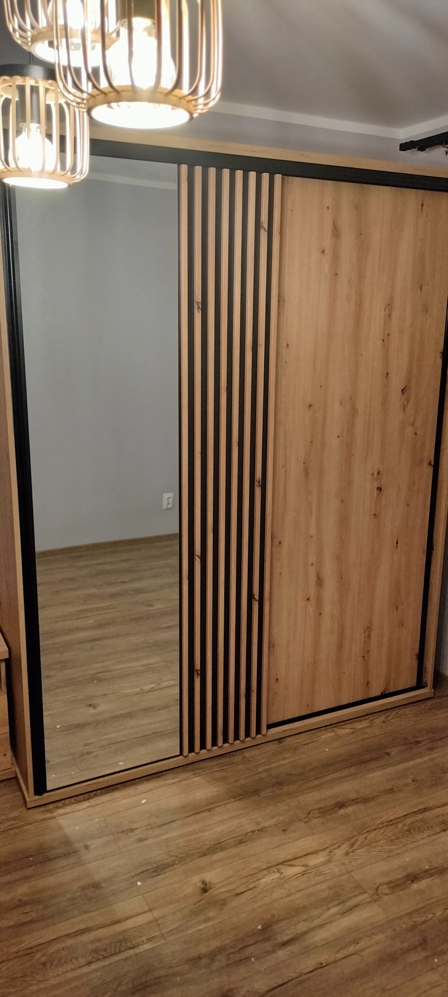 Duża, drewniana szafa przesuwna z lustrem i dekoracyjnymi listwami, widziana z bliskiej perspektywy, oświetlona przez wiszącą lampę z ażurowym kloszem. Widoczna część podłogi z paneli.