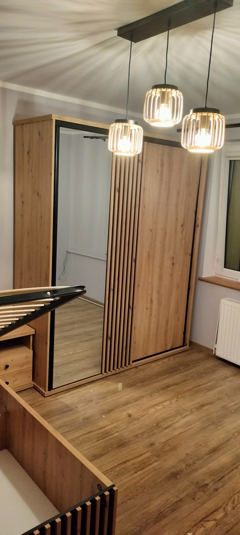 Szafa przesuwna z lustrem i drewnianymi lamelami w nowocześnie urządzonym pokoju, widoczne elementy łóżka w trakcie składania i wiszące lampy.