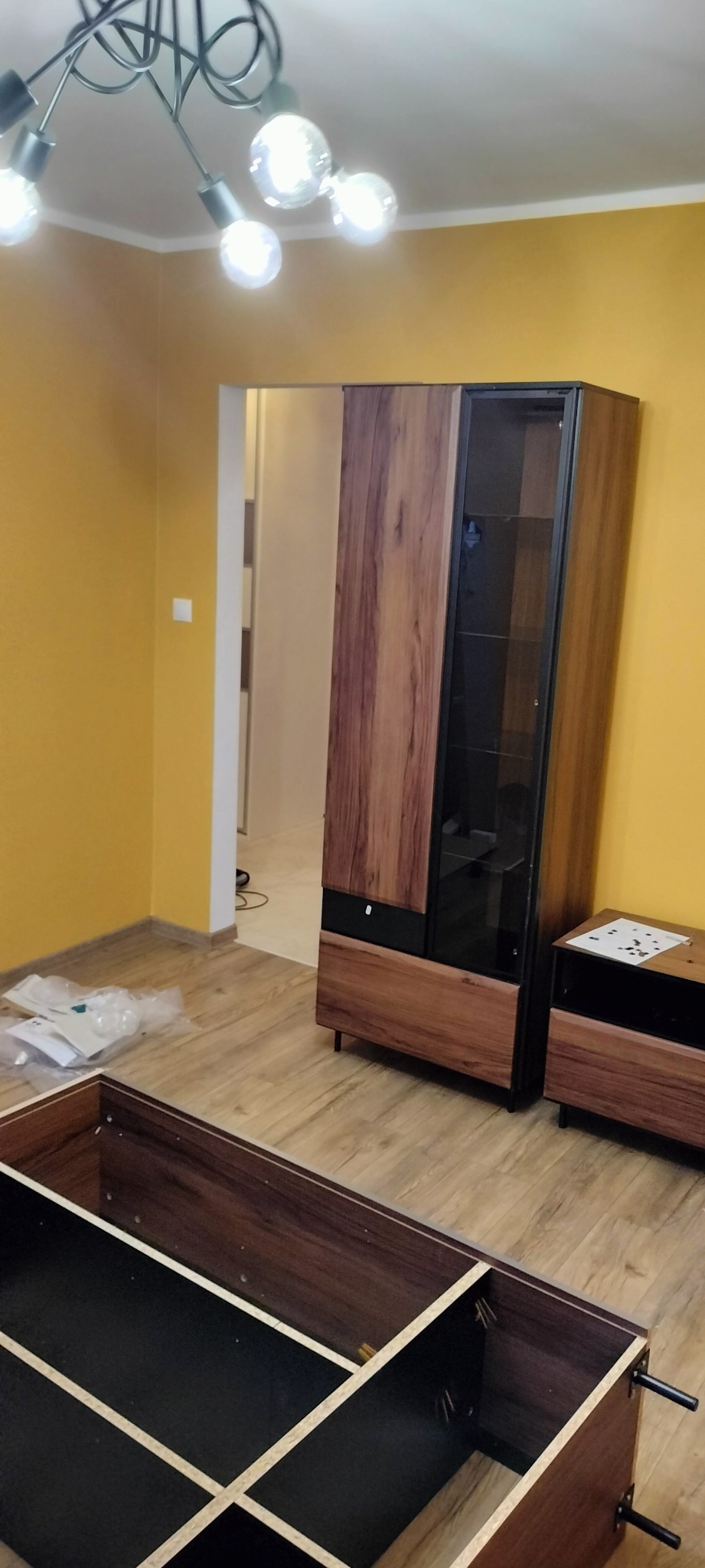 Fragment pokoju z żółtymi ścianami, gdzie widać szafę z przeszklonymi drzwiami i otwartą konstrukcję meblową w trakcie składania, z instrukcjami i elementami montażowymi leżącymi na podłodze.