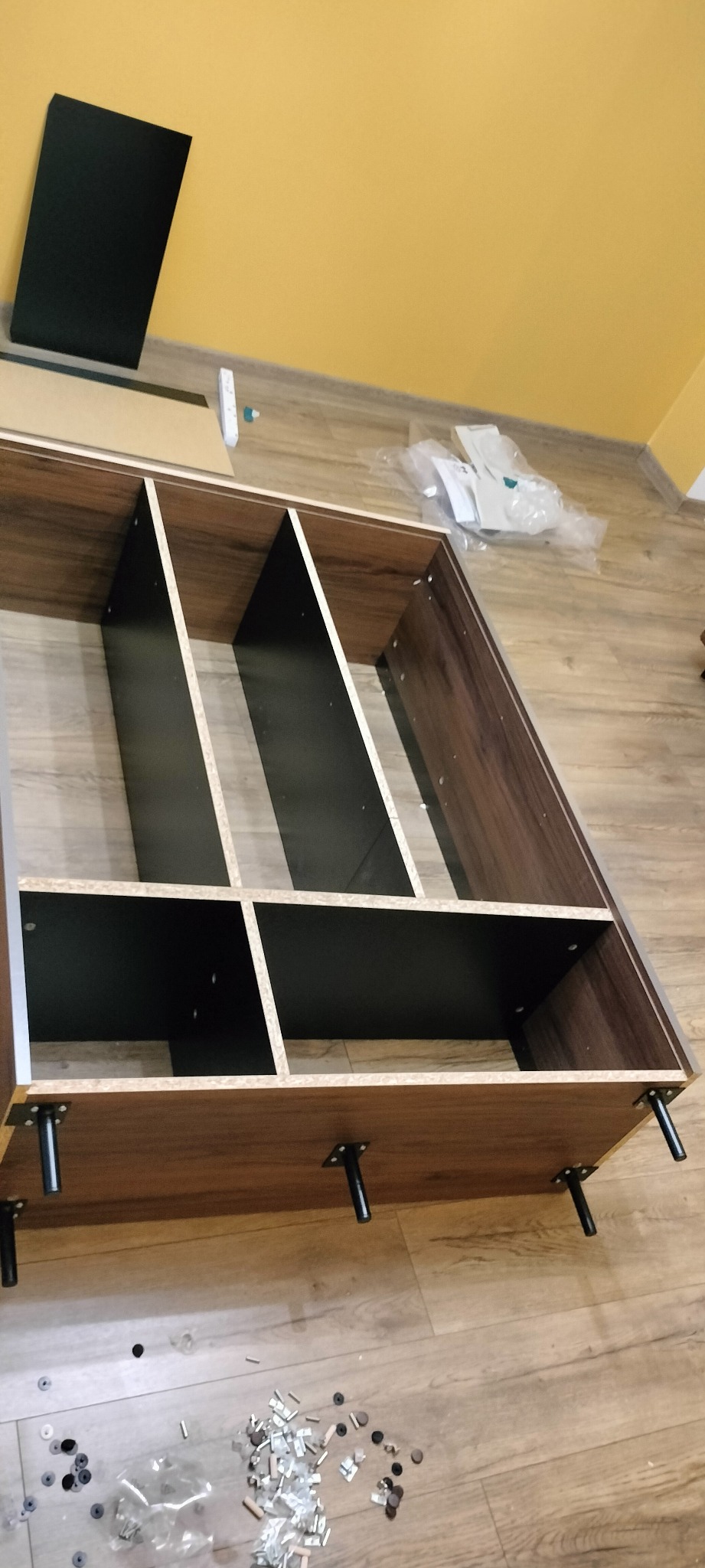 Półka z ciemnego drewna z czarnymi przegródkami w trakcie montażu, widoczne elementy montażowe rozsypane na podłodze.