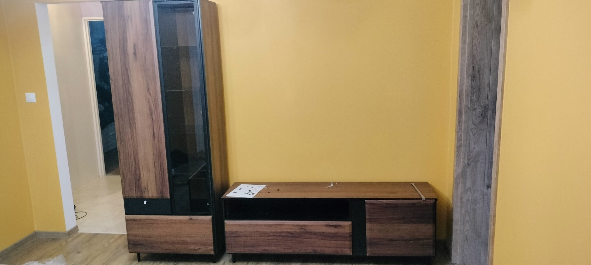 Nowoczesna szafka RTV z witryną i elementami w kolorze drewna na tle żółtej ściany, z widocznymi śladami po montażu w postaci papierów na blacie.