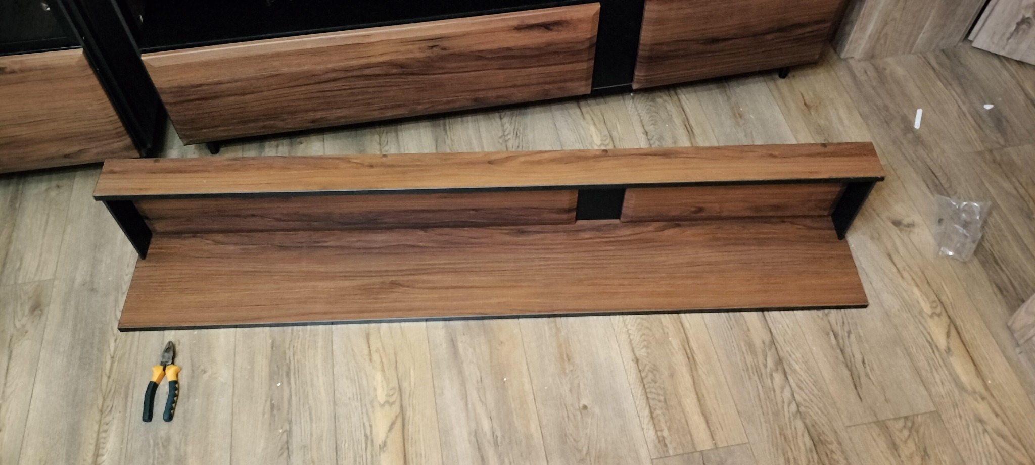 Brązowa półka wisząca z czarnymi elementami mocującymi, leżąca na panelach podłogowych, obok kombinerki z żółto-czarną rączką i folia z elementami montażowymi, prawdopodobnie w trakcie montażu.