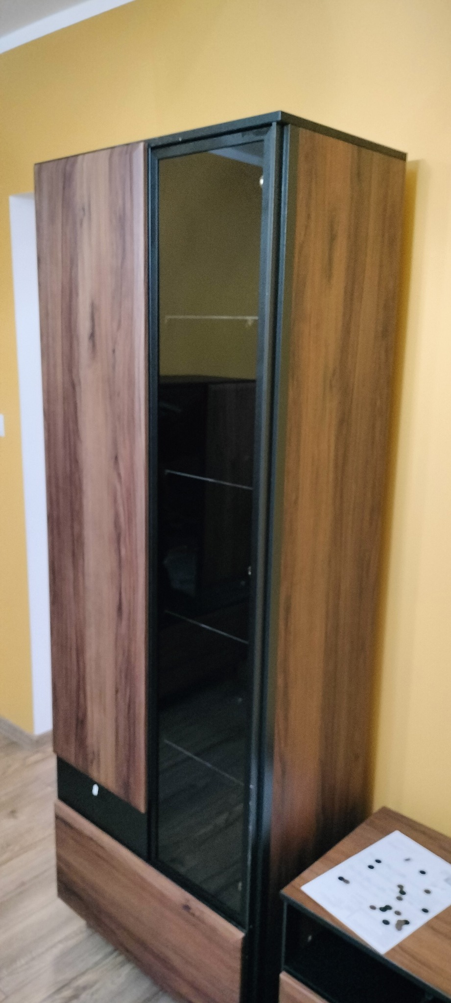 Wysoka szafa z drewnopodobnym wykończeniem, czarnymi ramami i szklanymi drzwiami, stojąca na tle żółtej ściany.