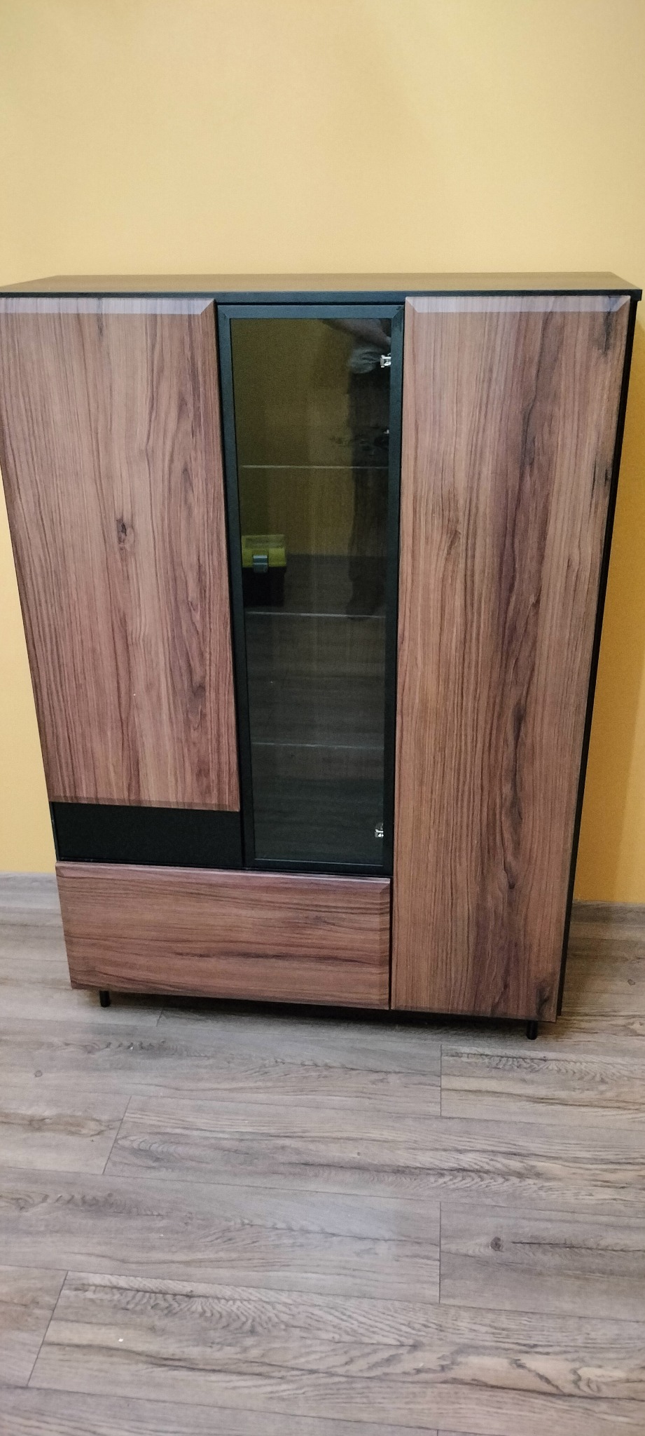 Nowoczesna komoda z drewnianymi frontami, czarnymi akcentami i przeszkloną witryną, stojąca na panelach podłogowych przed żółtą ścianą.