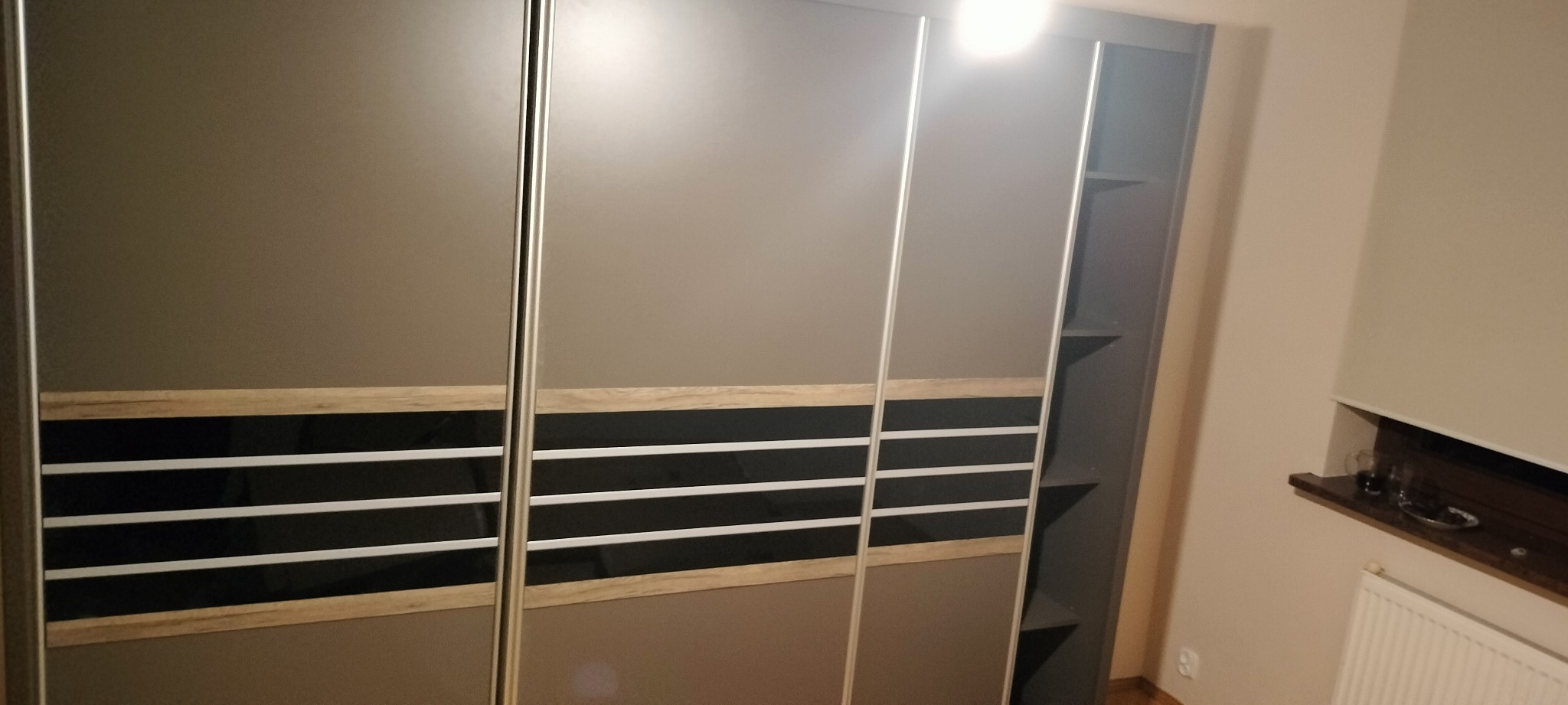 Szara szafa przesuwna z poziomymi zdobieniami w kolorze drewna i czerni, widoczne otwarte półki po prawej stronie szafy.