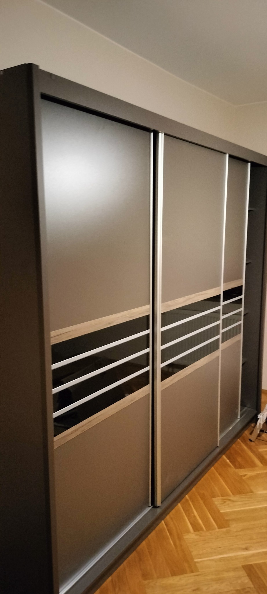 Nowoczesna szafa przesuwna z szarymi frontami, drewnianymi akcentami i poziomymi paskami, stojąca na parkiecie.