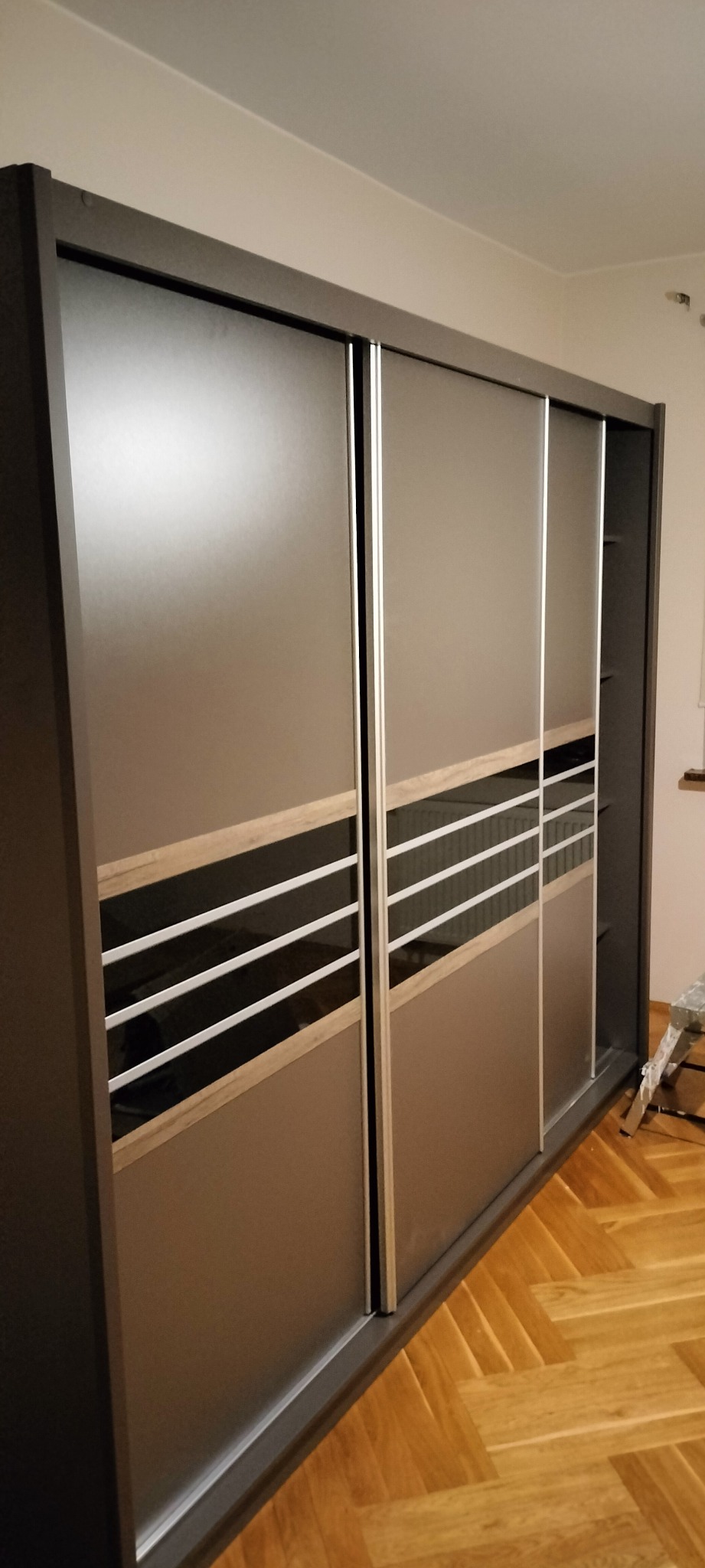 Nowoczesna szafa przesuwna z szarymi frontami, drewnianymi akcentami i poziomymi paskami, stojąca na parkiecie.