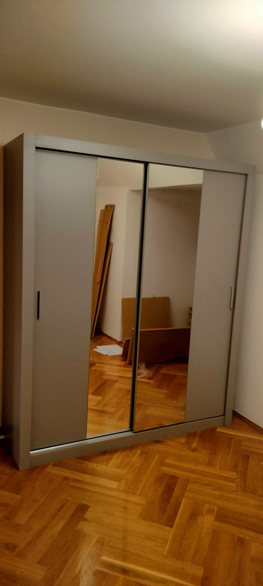 Szara szafa przesuwna z lustrami, odbijająca elementy wnętrza w trakcie montażu, widoczna na parkiecie.