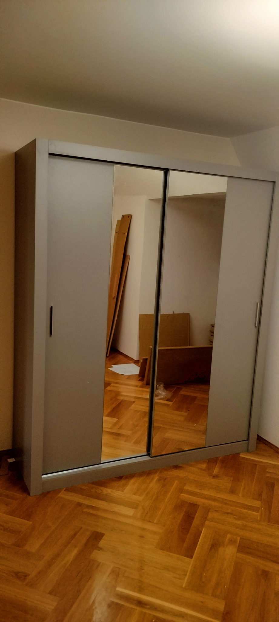 Szara szafa przesuwna z lustrami, widoczne płyty meblowe w odbiciu, na parkiecie ułożonym w jodełkę.