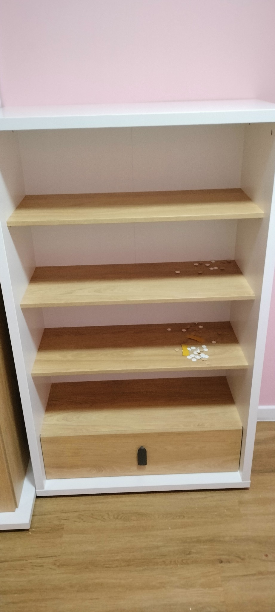 Biały regał z drewnianymi półkami i szufladą, na jednej z półek leżą białe zaślepki i kawałek żółtej taśmy, tło stanowi różowa ściana.