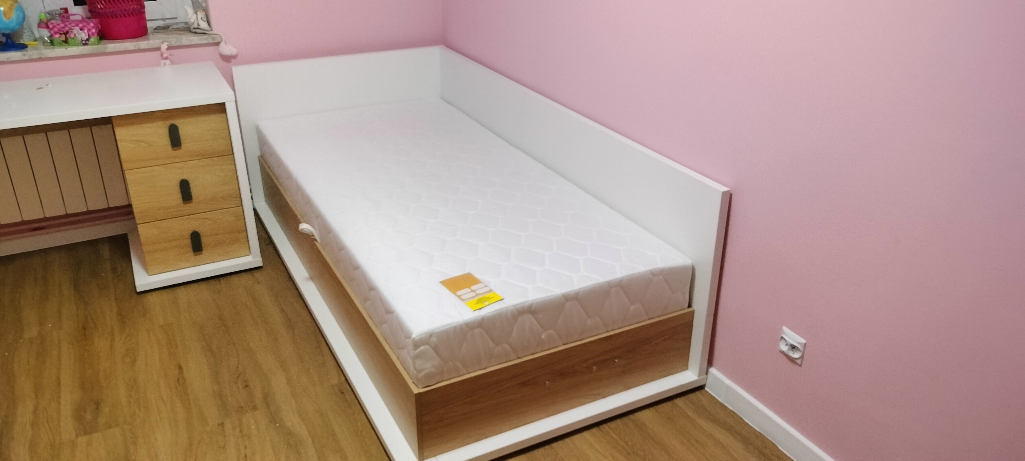 Nowoczesne łóżko dziecięce z białymi bokami i drewnianą ramą, materac z metką, obok biurko z szufladami, różowe ściany w tle.