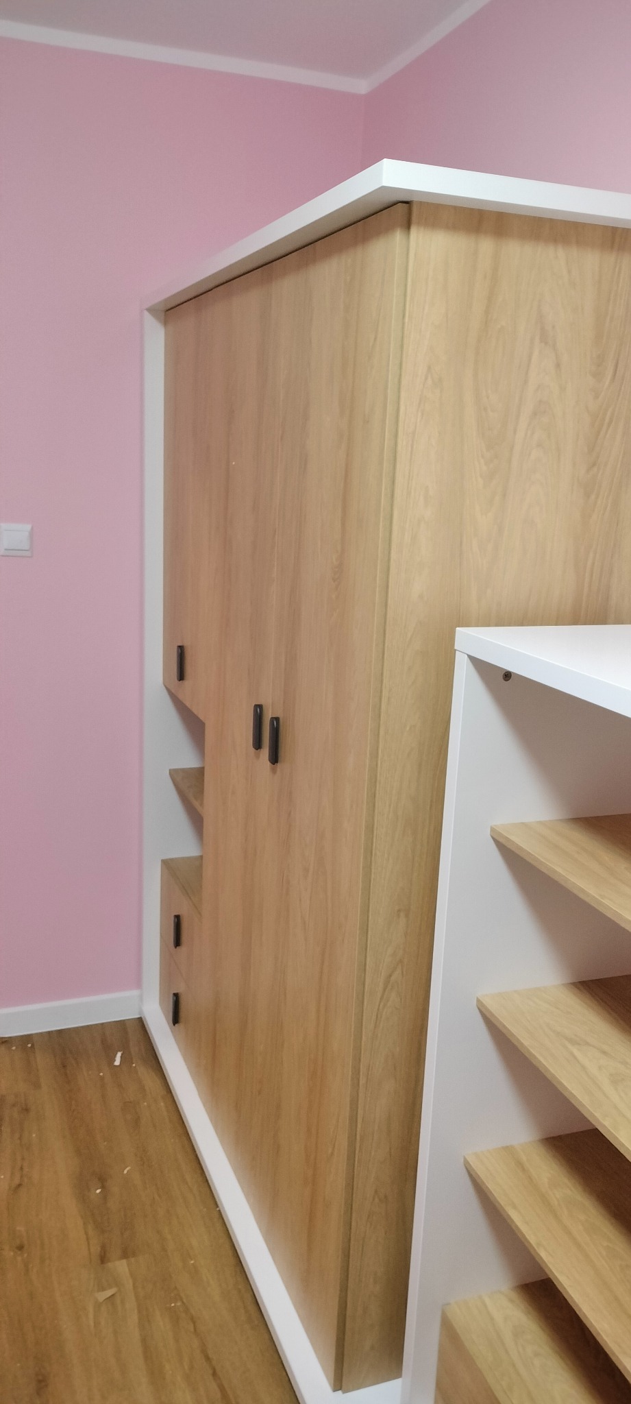 Wbudowana szafa z drewnianymi frontami i czarnymi uchwytami, z otwartymi półkami po prawej stronie, na tle różowej ściany.
