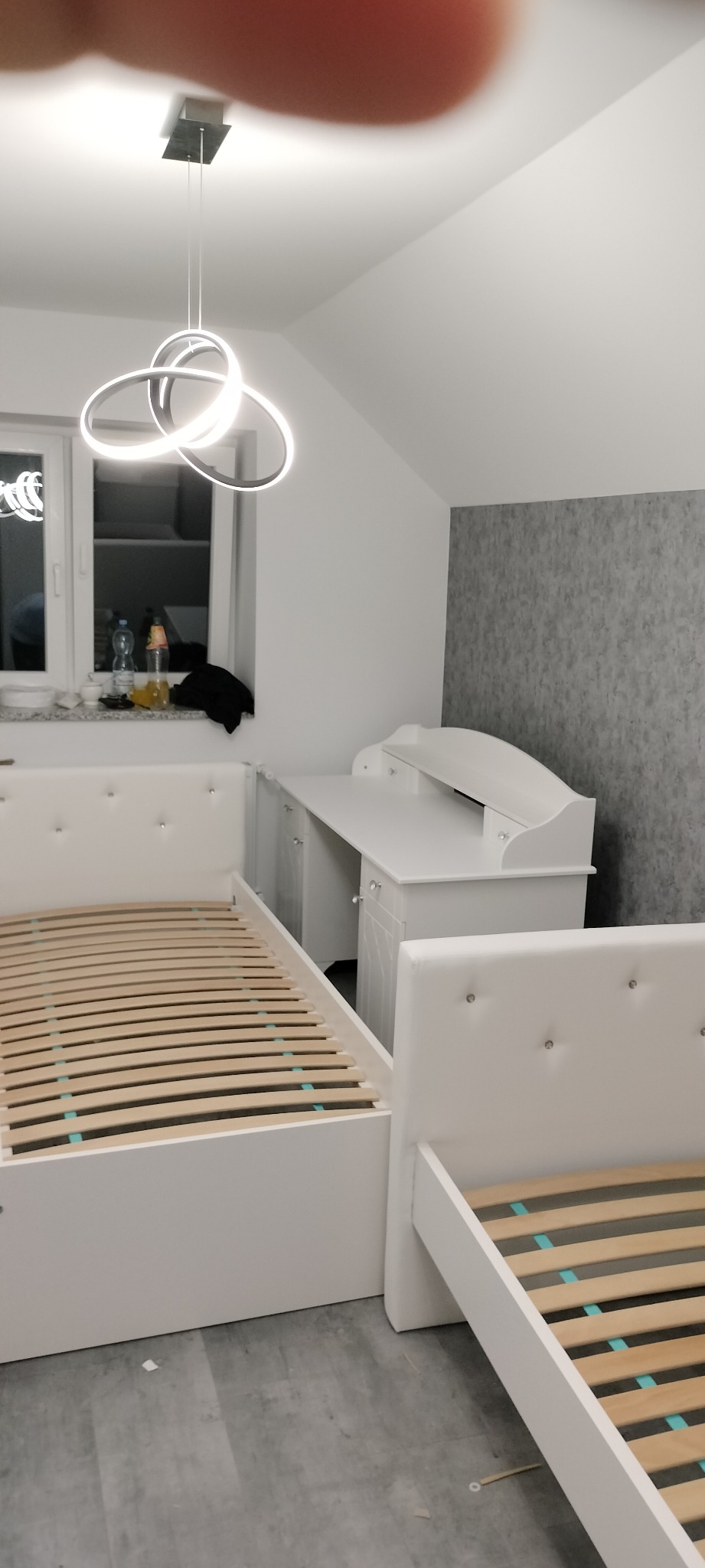 Pokój dziecięcy z dwoma białymi łóżkami z tapicerowanymi zagłówkami ozdobionymi kryształkami oraz białym biurkiem z nadstawką, oświetlony nowoczesną lampą wiszącą.