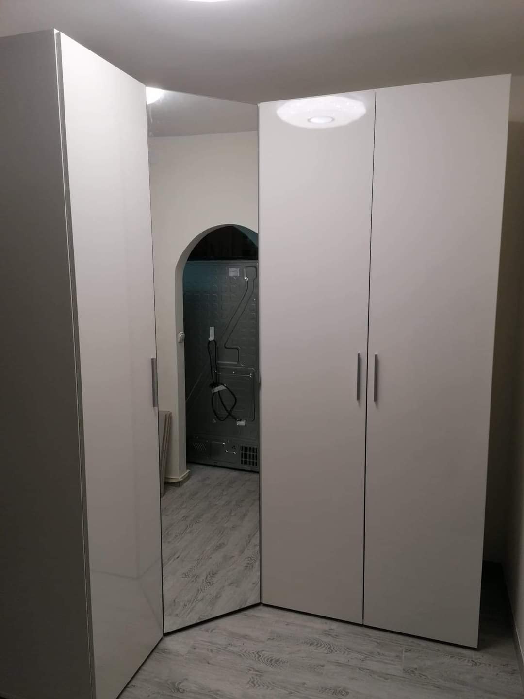 Wnętrze z narożną, białą szafą z połyskiem, widokiem na pralkę przez łukowe przejście i szare panele podłogowe.