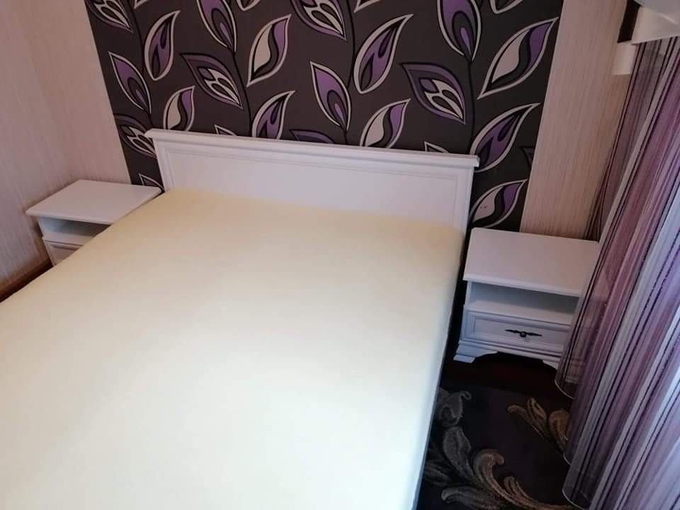 Sypialnia z białymi meblami: łóżko z dwoma szafkami nocnymi, ściana z wzorzystą tapetą w odcieniach szarości i fioletu, zasłona w paski.