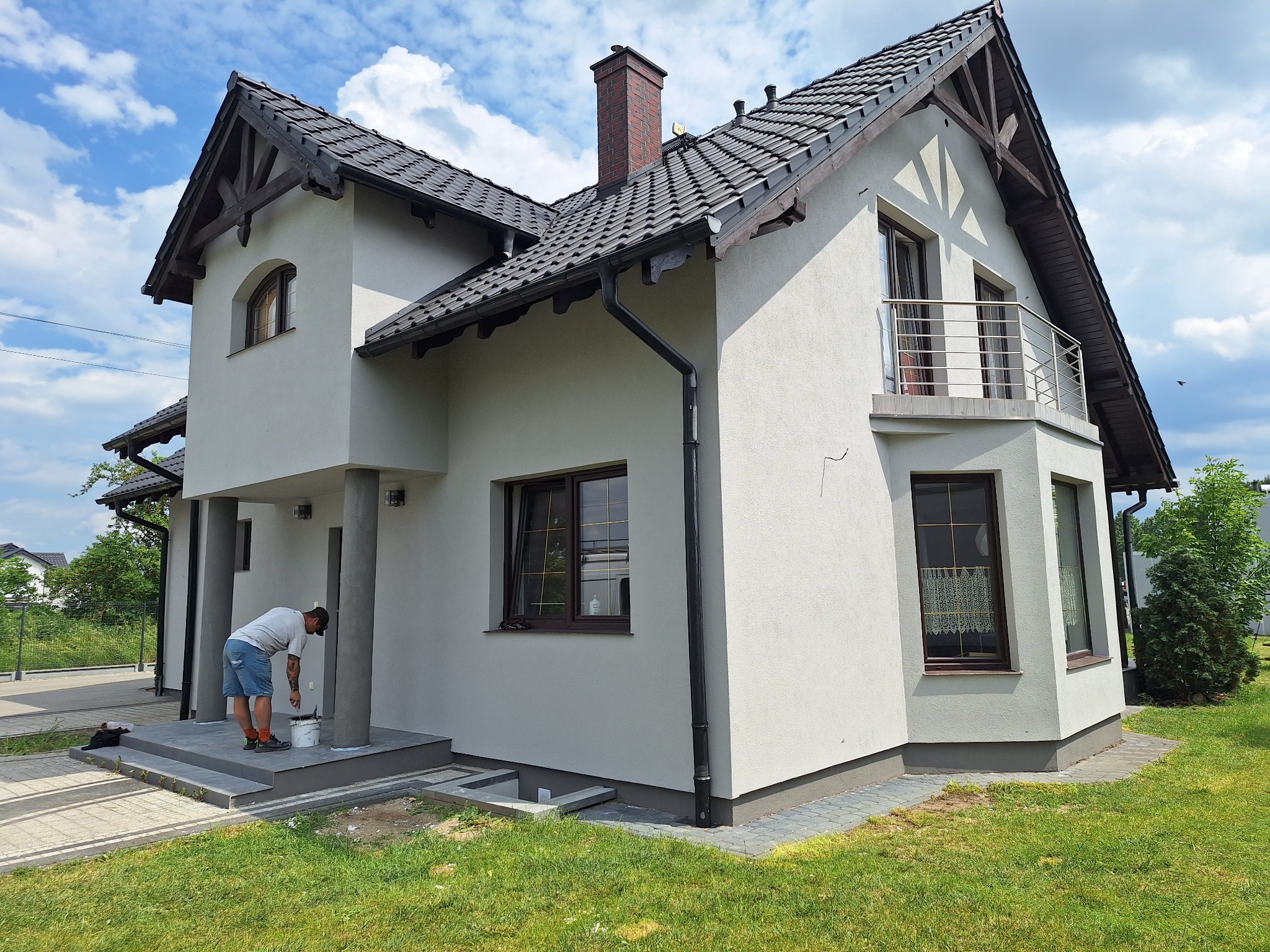 Dwukondygnacyjny dom z tynkowaną, jasną elewacją. Mężczyzna maluje filar wejściowy. Widoczne detale architektoniczne i fragment ogrodu.