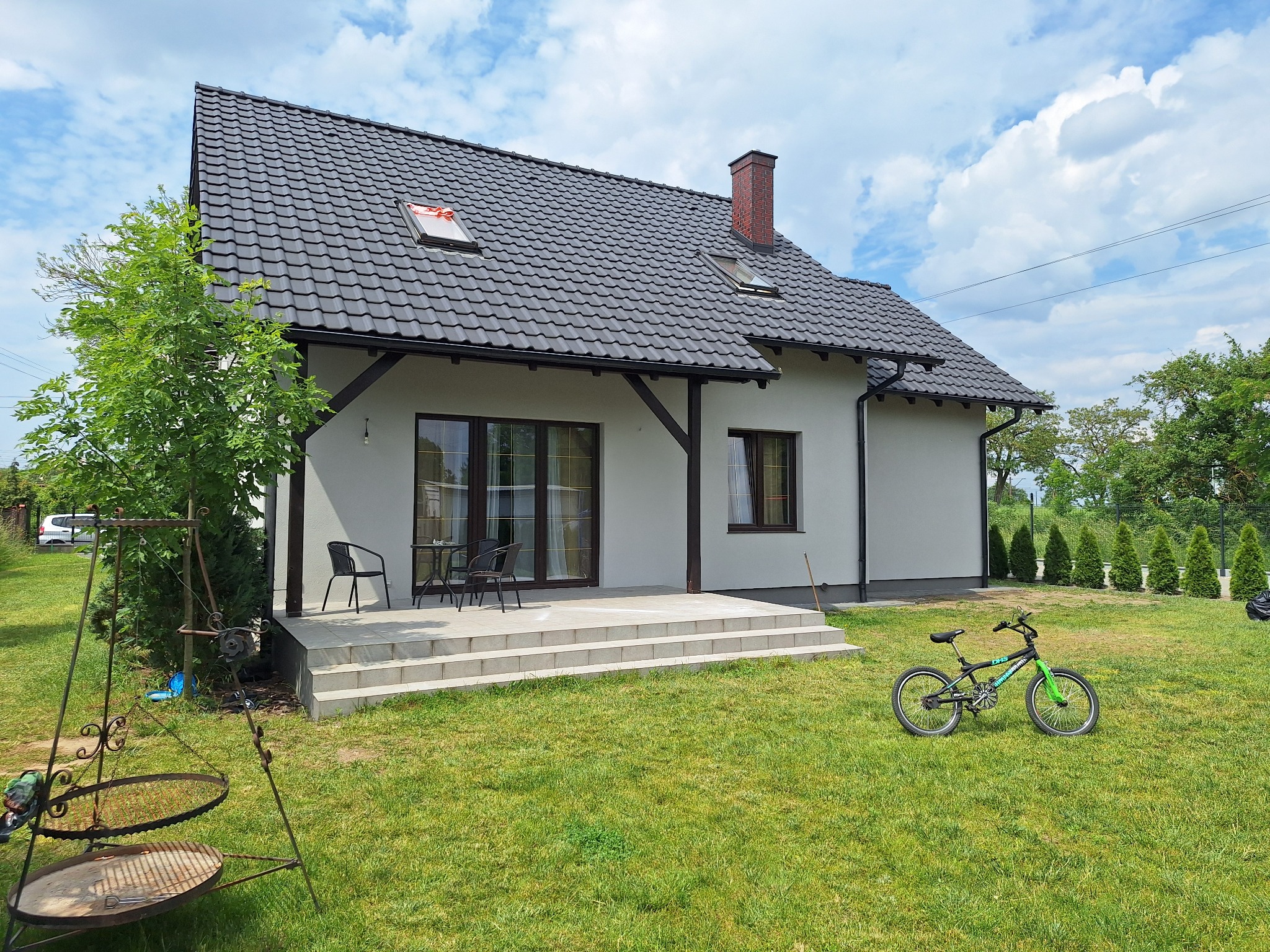 Jasny dom z ciemną dachówką i nową elewacją, widok z ogrodu. Na trawniku stoi rower. Widoczne okna z brązowymi ramami i schody prowadzące do drzwi.