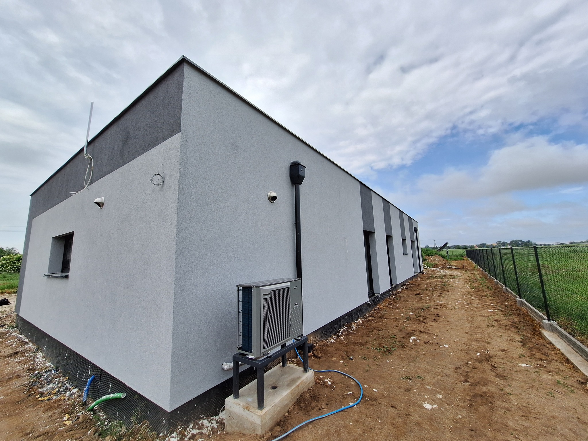 Budynek z szarą elewacją i ciemniejszymi akcentami, widok z zewnątrz, z zainstalowanym klimatyzatorem na fundamencie. Tło: niebo i ogrodzenie.