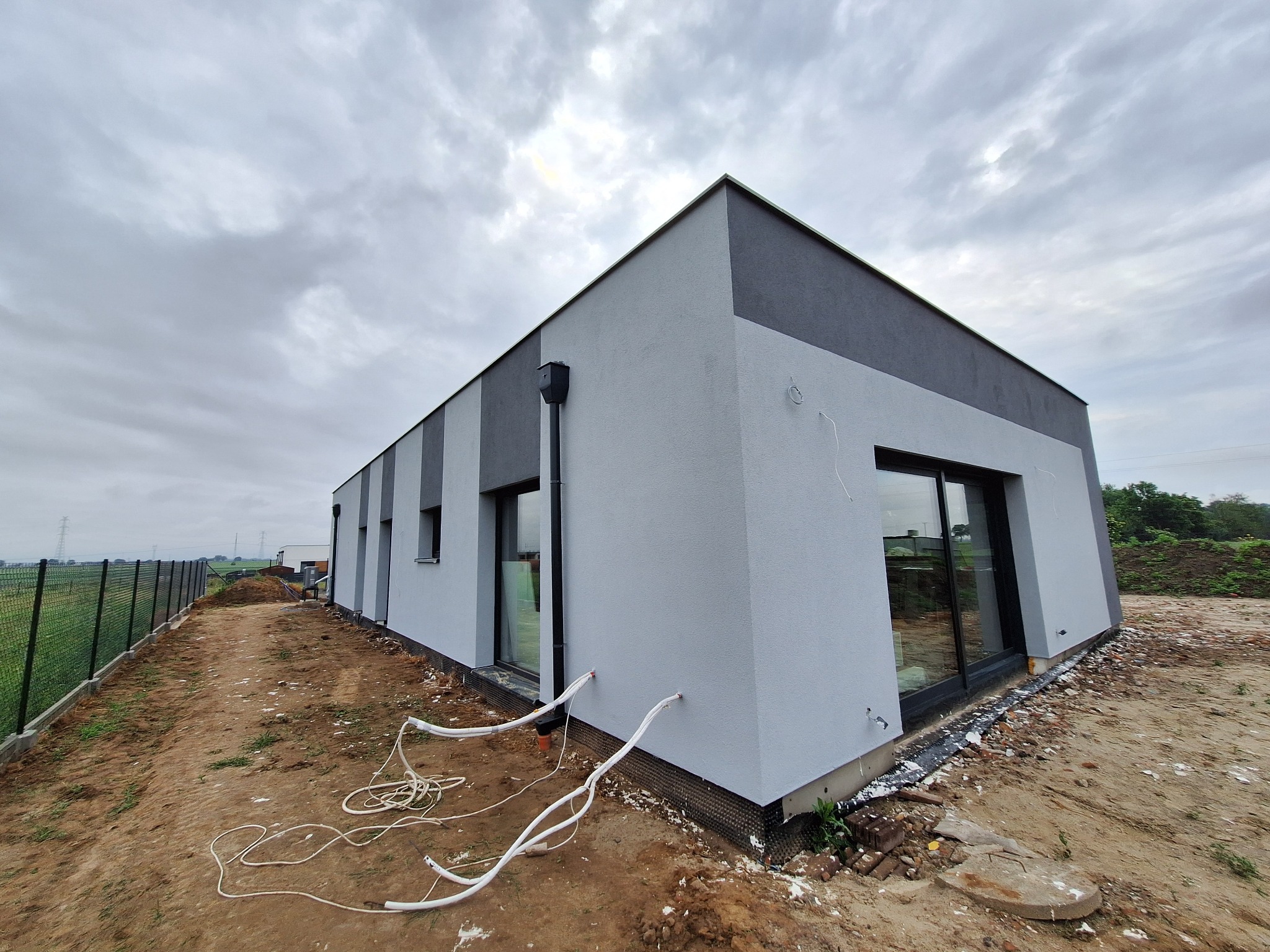 Nowoczesny dom z szarą elewacją i czarnymi oknami, widok z zewnątrz, teren wokół w trakcie zagospodarowania, pochmurne niebo.