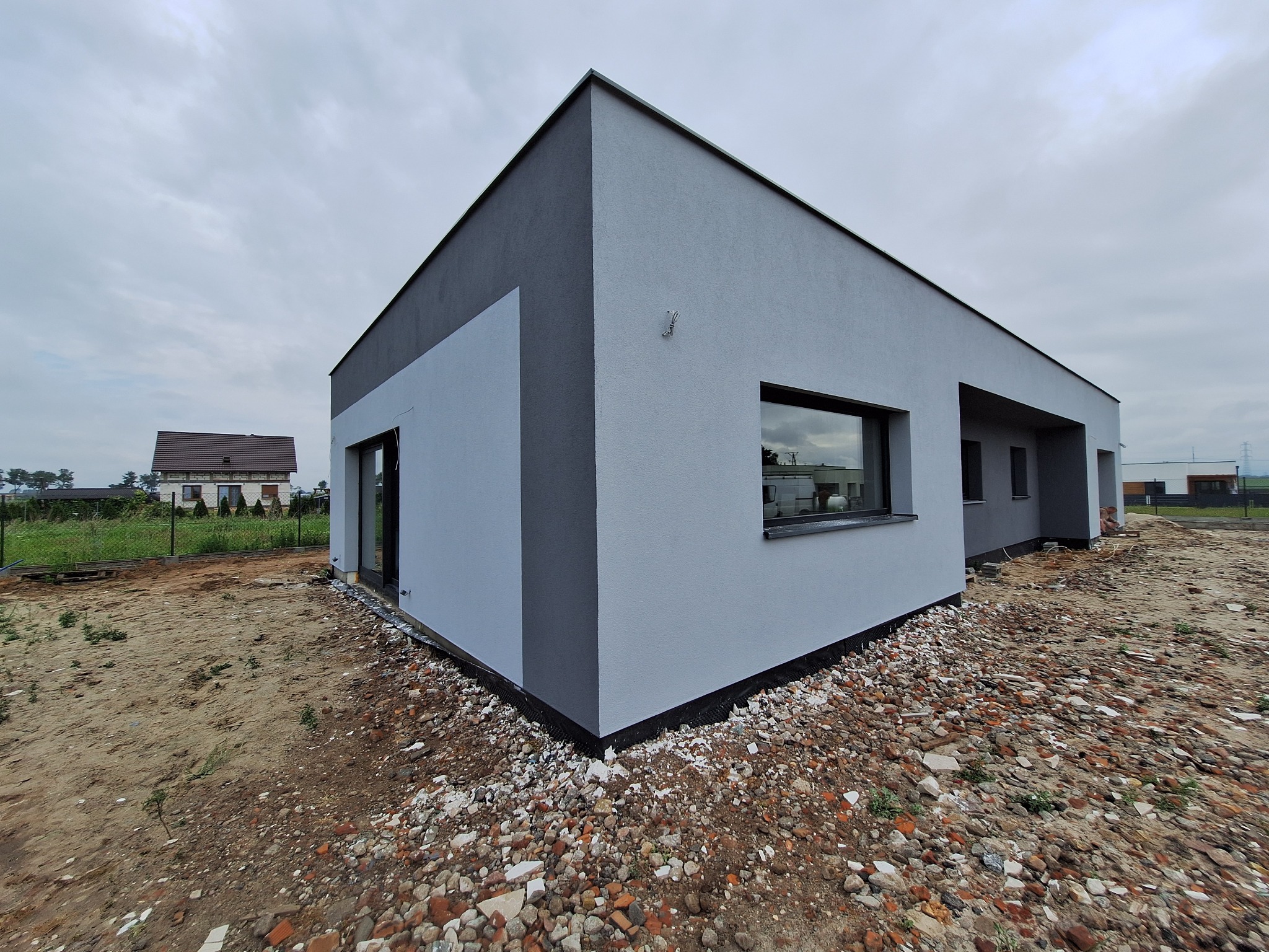 Nowoczesny, parterowy dom z elewacją w odcieniach szarości, z widocznym fragmentem dachu i okna. W tle inny budynek mieszkalny. Działka w trakcie zagospodarowywania.