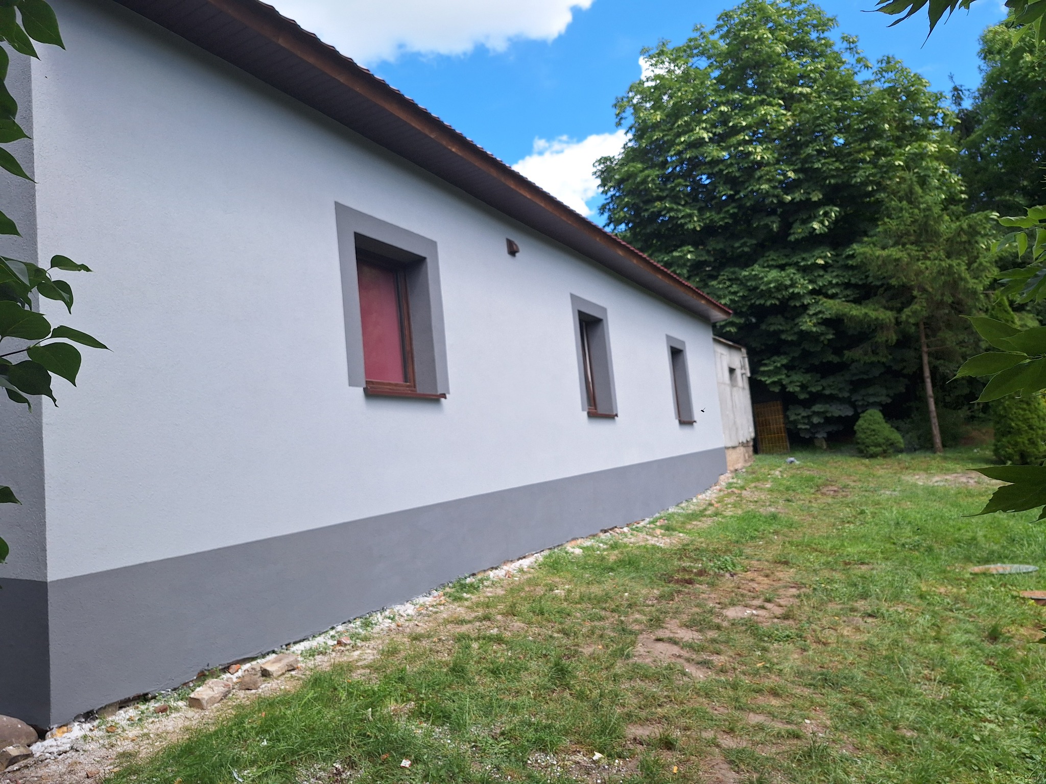 Ocieplony budynek z cegły, pomalowany na szaro z ciemniejszym pasem u dołu, okna w szarych ramach, widok z boku na tle zieleni i błękitnego nieba.