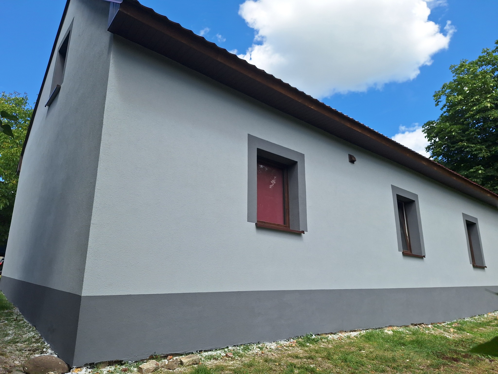 Odnowiona elewacja budynku po ociepleniu i pomalowaniu na szaro, z widoczną podbitką dachową i oknami w ciemnych ramach. Część elewacji pomalowana na ciemniejszy odcień szarości.