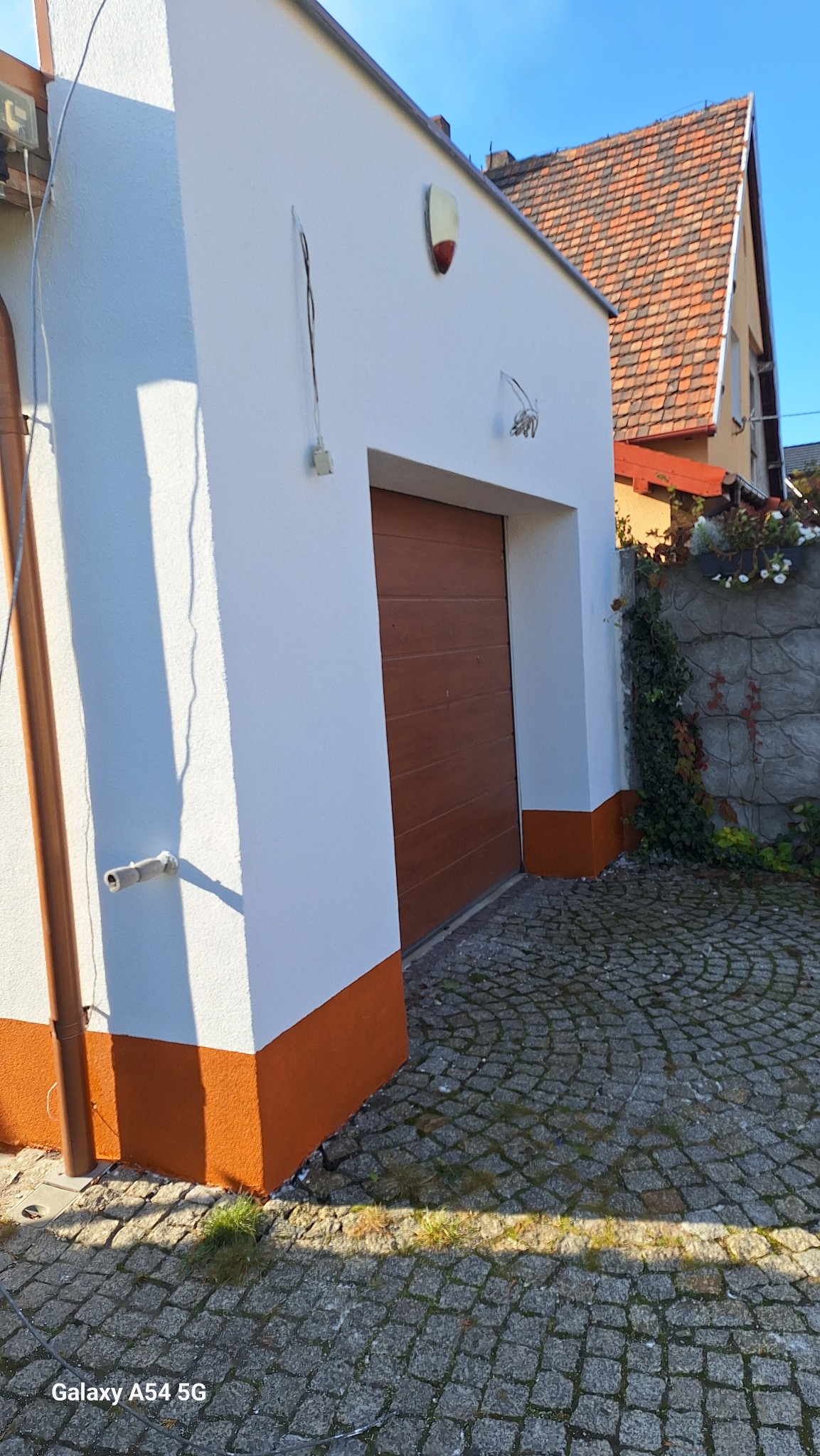 Odnowiona elewacja budynku z brązowymi akcentami u dołu i wokół brązowej bramy garażowej, na tle kostki brukowej i fragmentu domu sąsiada.