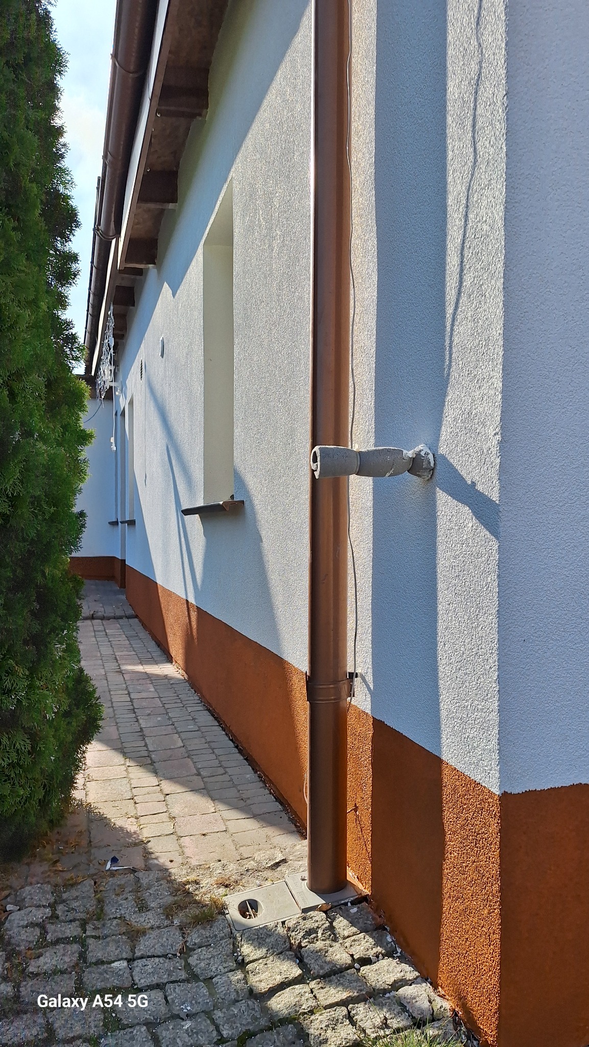 Elewacja budynku z tynkiem barankiem, pomalowana na biało z brązowym cokołem. Widoczna rynna i chodnik z kostki brukowej. Jasne, słoneczne oświetlenie.