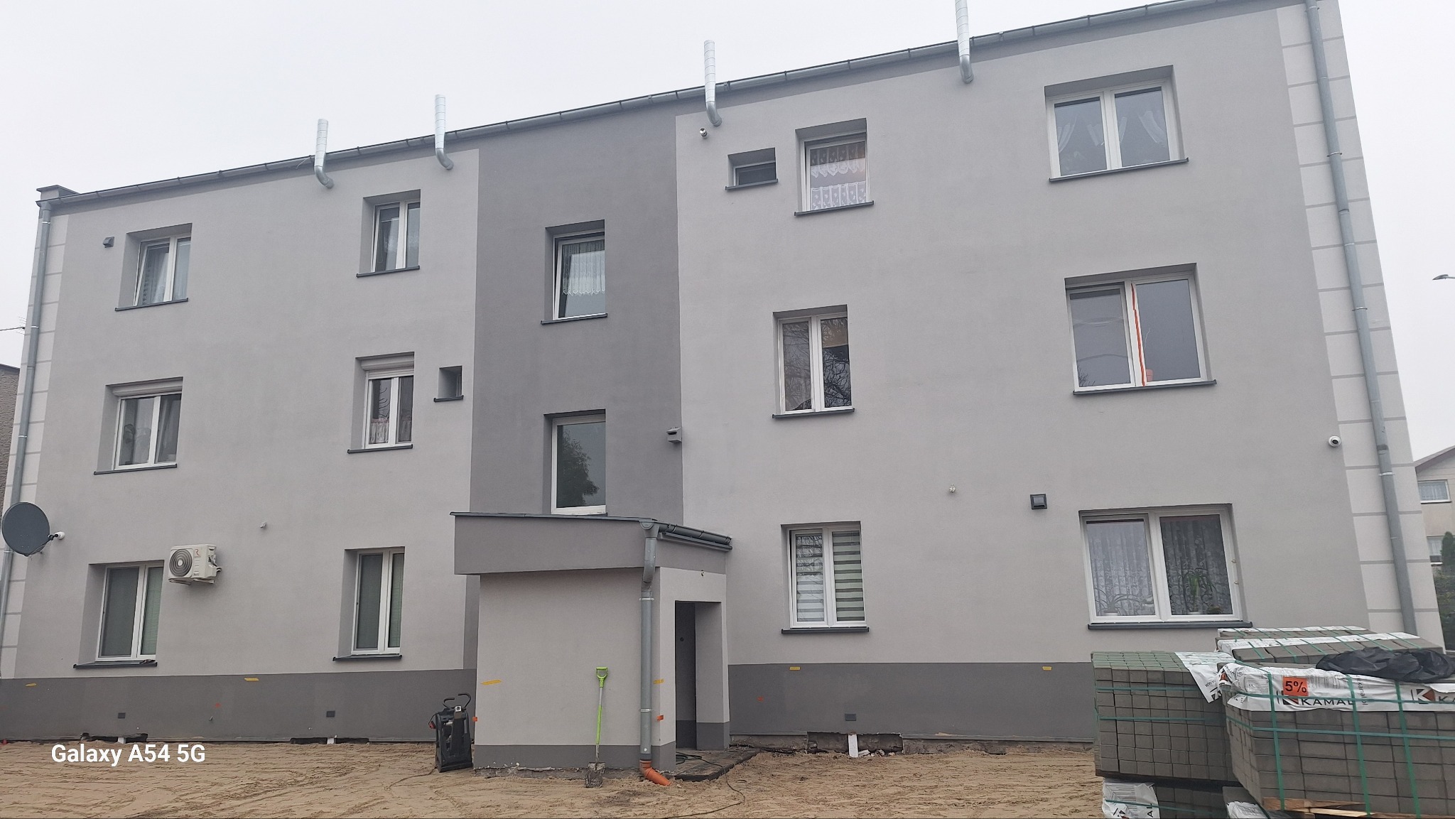 Ocieplona kamienica z nową, jasnoszarą elewacją i ciemniejszym cokołem. Widoczne kominy wentylacyjne, klimatyzator, ułożona kostka brukowa.