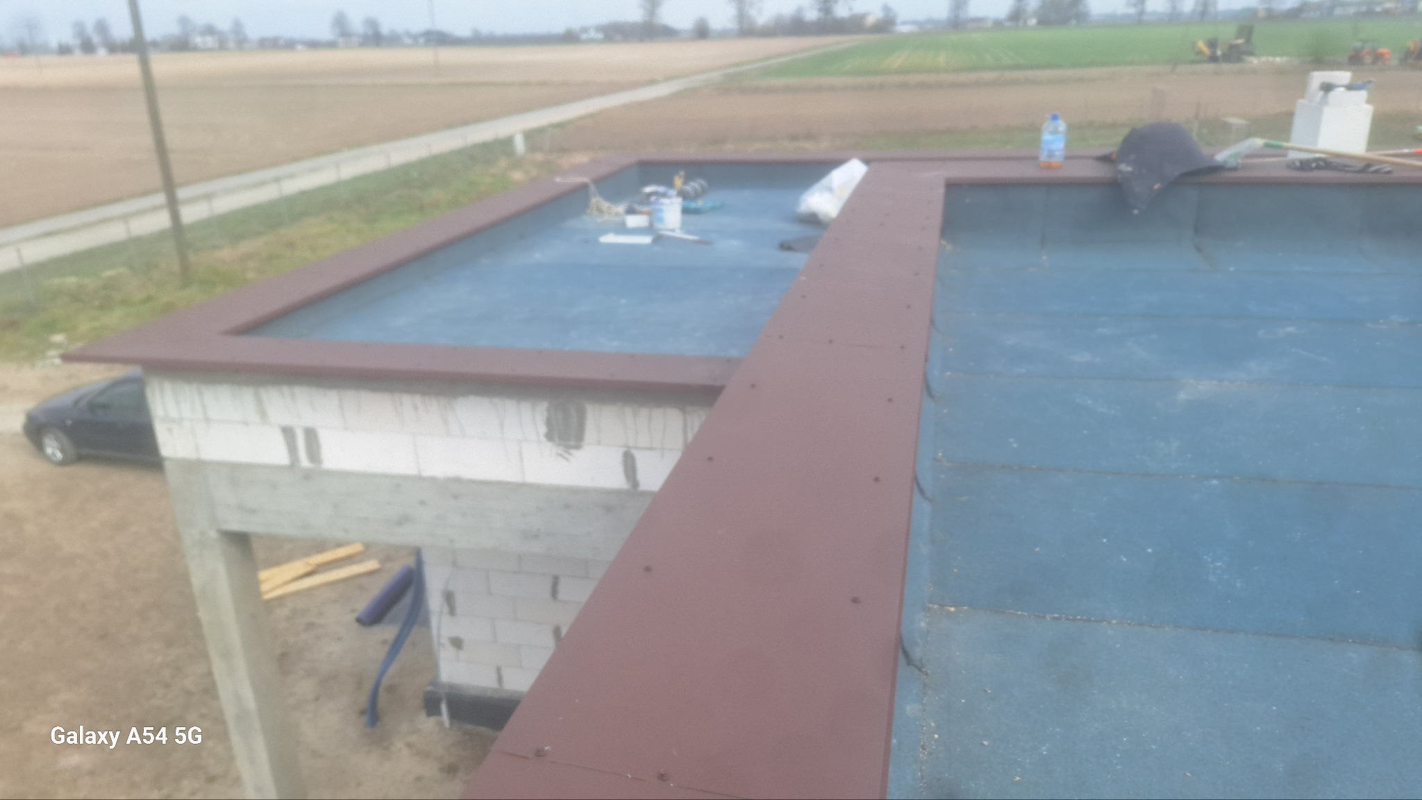 Płaski dach budynku w trakcie hydroizolacji papą, widoczne narzędzia i materiały budowlane. W tle krajobraz polny i fragment samochodu.