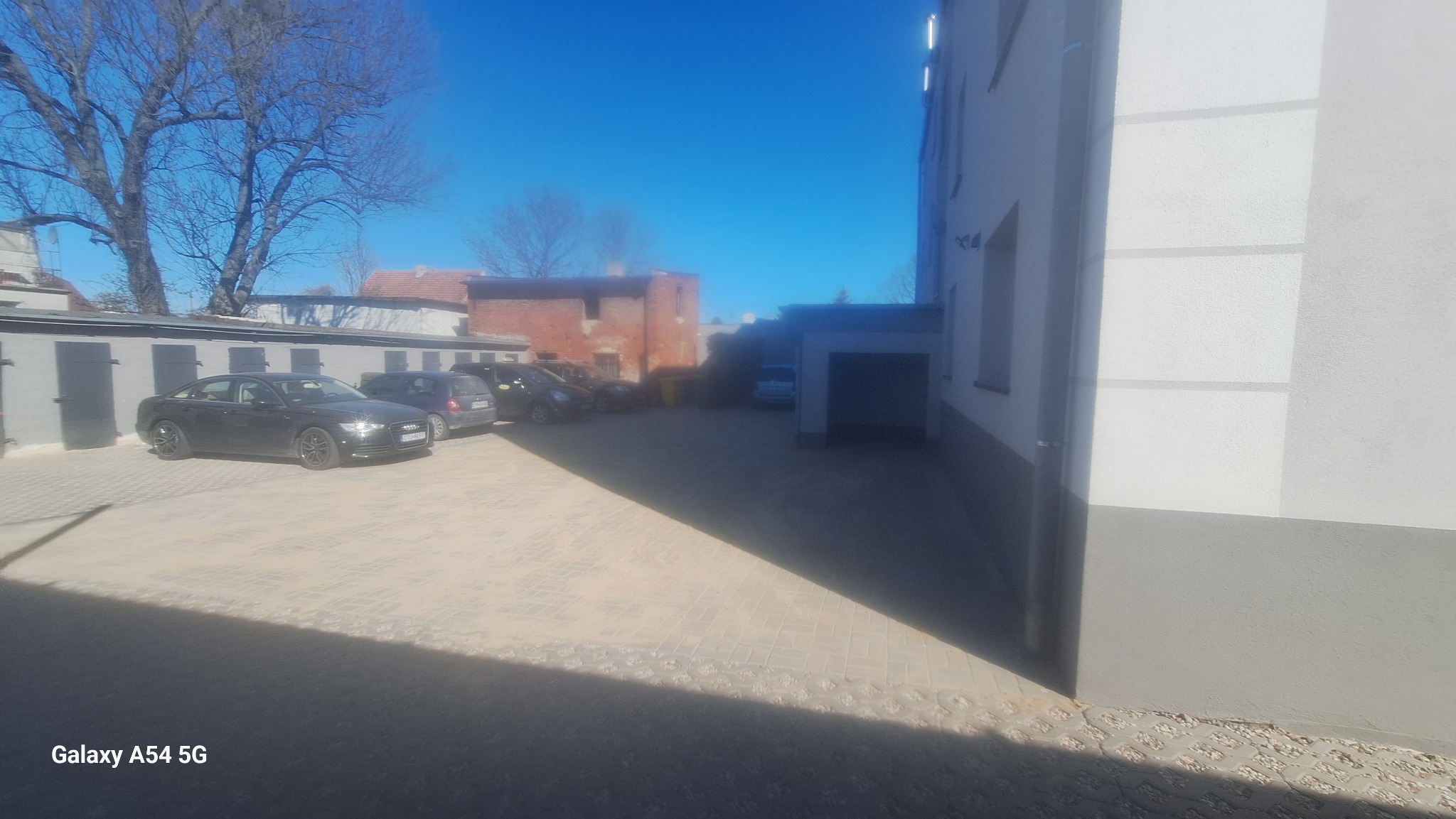 Elewacja kamienicy po renowacji z ociepleniem, widoczna z poziomu podwórka z kostką brukową i zaparkowanymi samochodami. Jasna elewacja, szara podmurówka.