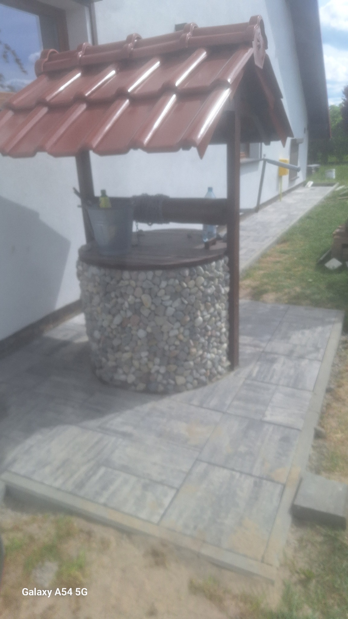 Studnia ozdobna z kamienną obudową i czerwoną dachówką, otoczona szarą kostką brukową. Tło stanowi biały budynek z oknem. Ujęcie z dołu, lekko z lewej strony.