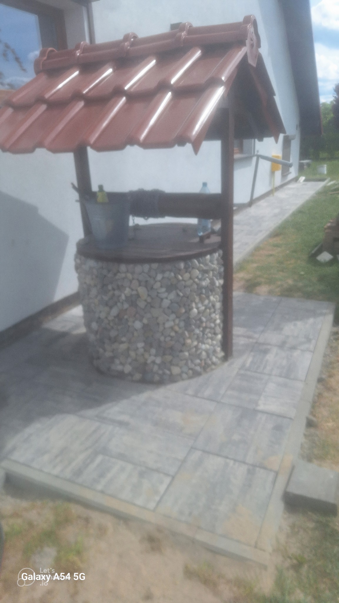 Studnia ogrodowa z kamienną obudową i brązowym dachem z dachówki, ustawiona na szarym brukowanym podłożu przy białym budynku.