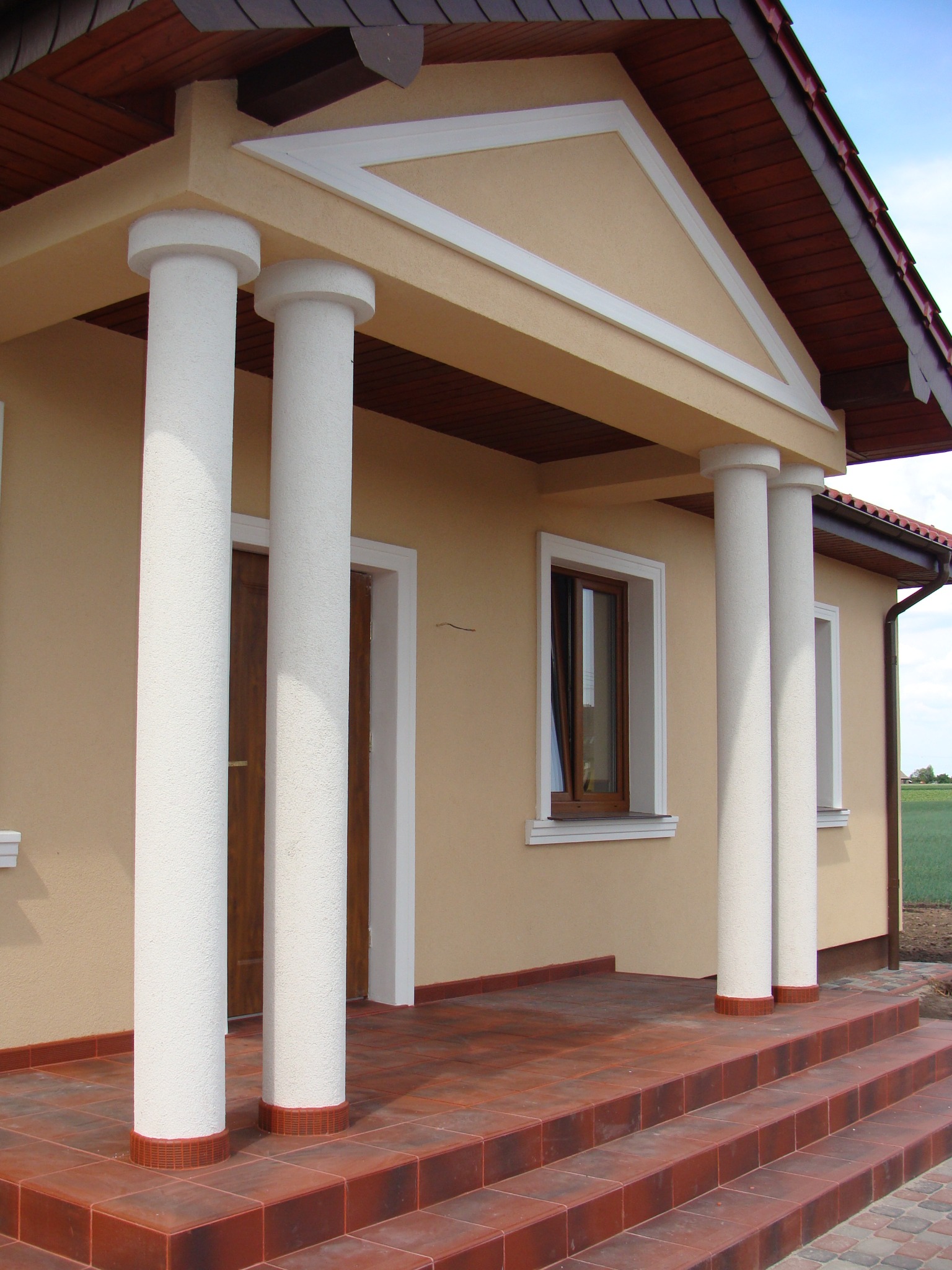 Ocieplony budynek z beżową elewacją, białymi kolumnami i schodami wyłożonymi czerwoną terakotą. Widok na fragment budynku z wejściem.