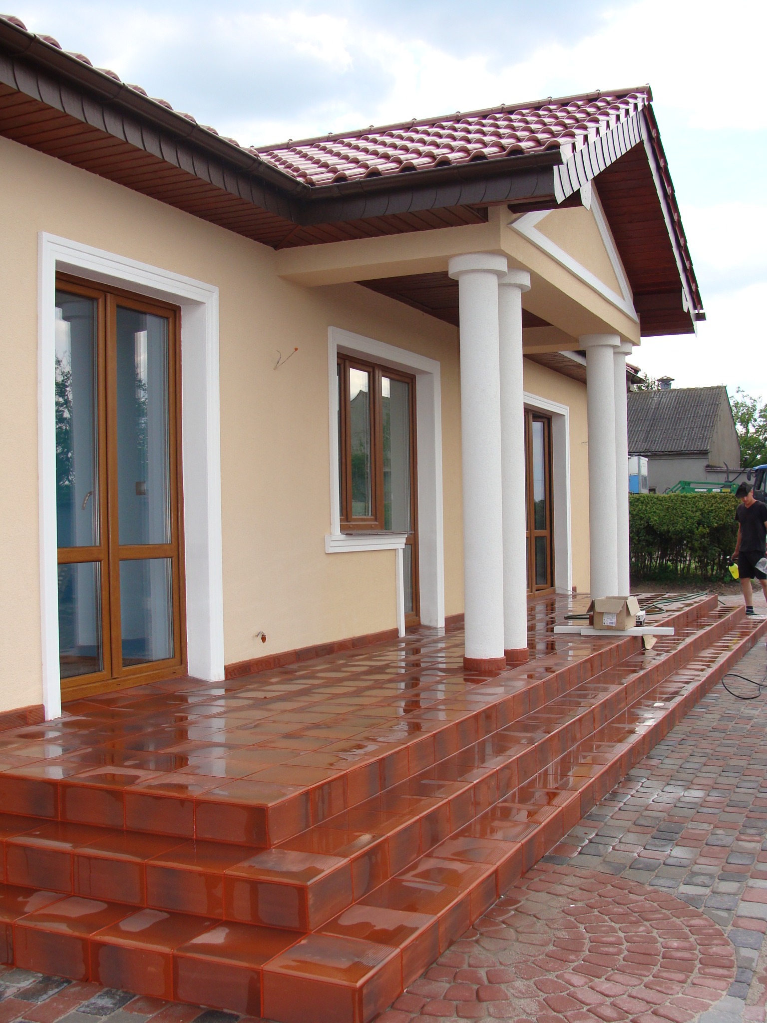 Ocieplony dom z kolumnami i schodami wyłożonymi błyszczącymi, brązowymi płytkami. Widoczna elewacja w odcieniach beżu i brązu oraz fragment posesji.