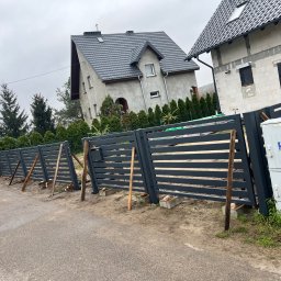 ESMET - Montaż nowoczesnego, poziomego ogrodzenia z metalowych paneli w kolorze grafitowym, widoczne drewniane podpory stabilizujące konstrukcję w trakcie instalacji, w tle dom w stanie surowym.