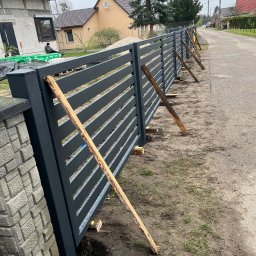 ESMET - Montaż nowoczesnego ogrodzenia panelowego w kolorze grafitowym, tymczasowo podpartego drewnianymi stemplami, widoczne słupki ogrodzeniowe i fragment drogi.