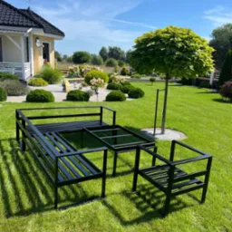 Stelaż metalowy mebli ogrodowych w kolorze czarnym, składający się z sofy, stołu i krzesła, ustawiony na trawniku przed domem z zadbanym ogrodem w słoneczny dzień.