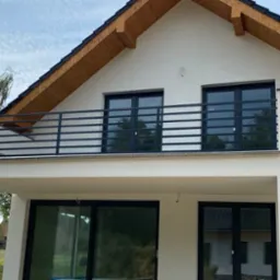 Nowoczesna, pozioma, metalowa balustrada balkonowa w kolorze antracytowym zamontowana na białym domu z drewnianym okapem dachowym. Widok na zielone drzewa odbijające się w szybach okiennych.