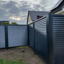 ESMET - Nowoczesne, poziome, szare ogrodzenie lamelowe z metalu, widok z bliska na narożnik, z fragmentem domu w tle i pochmurnym niebem.