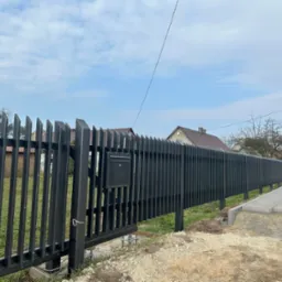 Nowoczesne, czarne ogrodzenie z pionowych, metalowych profili z bramą i skrzynką na listy, widoczne ślady prac ziemnych przy chodniku.