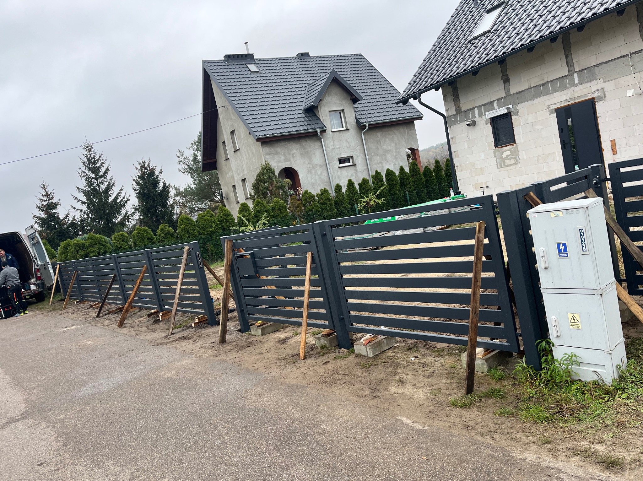 Montaż nowoczesnego, poziomego ogrodzenia z metalowych paneli w kolorze grafitowym, widoczne drewniane podpory stabilizujące konstrukcję w trakcie instalacji, w tle dom w stanie surowym.