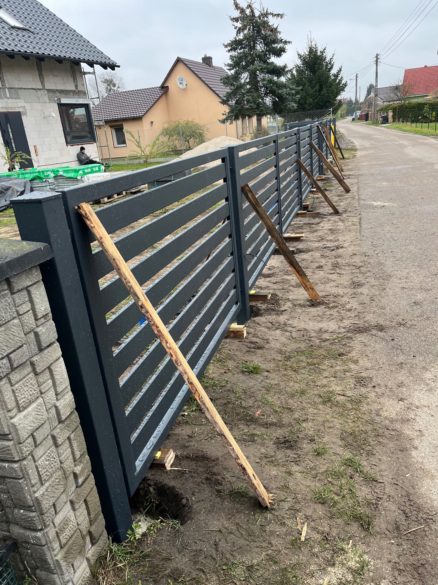 Montaż nowoczesnego ogrodzenia panelowego w kolorze grafitowym, tymczasowo podpartego drewnianymi stemplami, widoczne słupki ogrodzeniowe i fragment drogi.