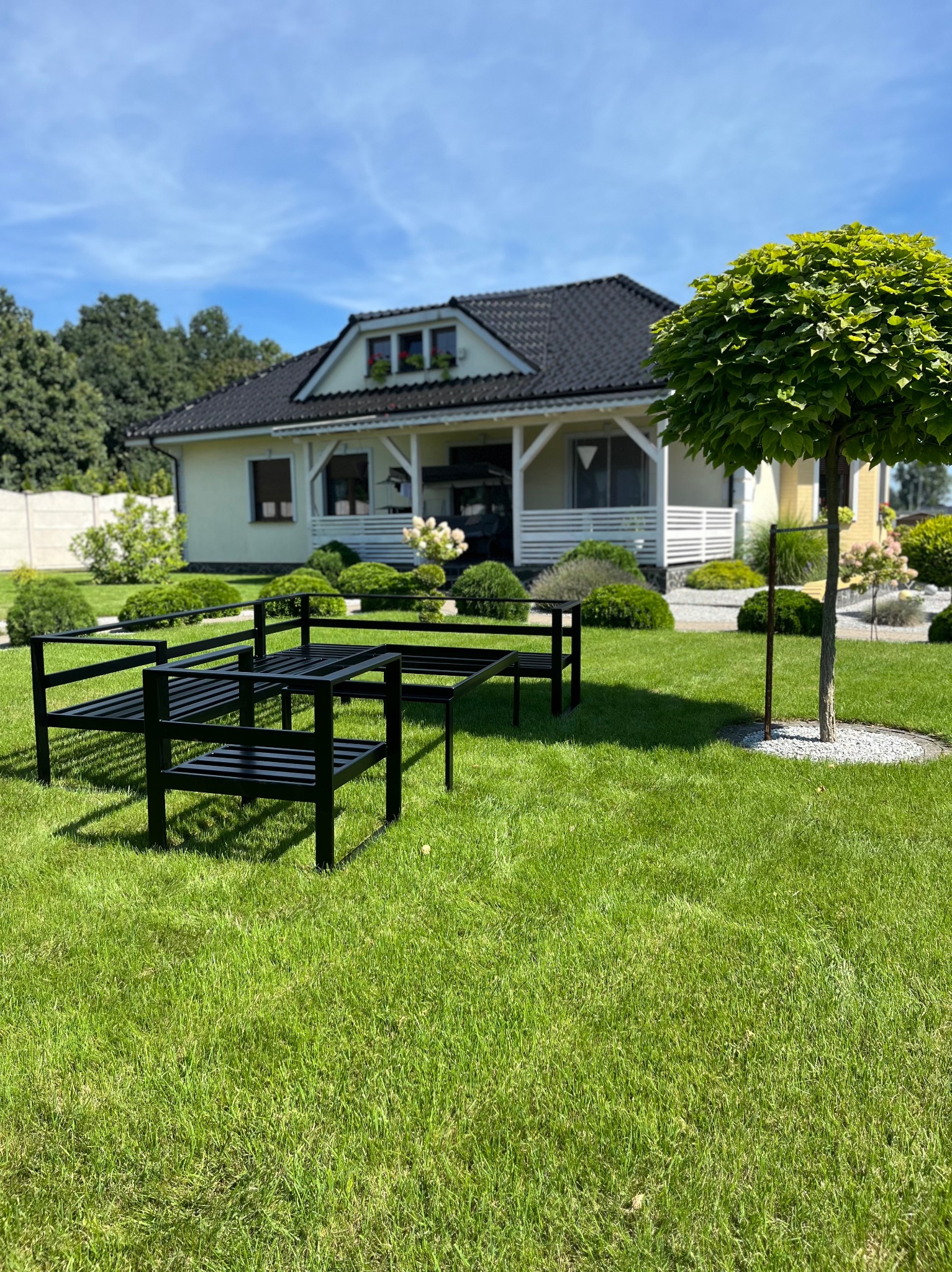 Czarny, metalowy stelaż pod przyszłe ogrodzenie posesji, widoczny na tle zadbanego trawnika, domu z białymi elementami i dekoracyjnymi krzewami, w słoneczny dzień.