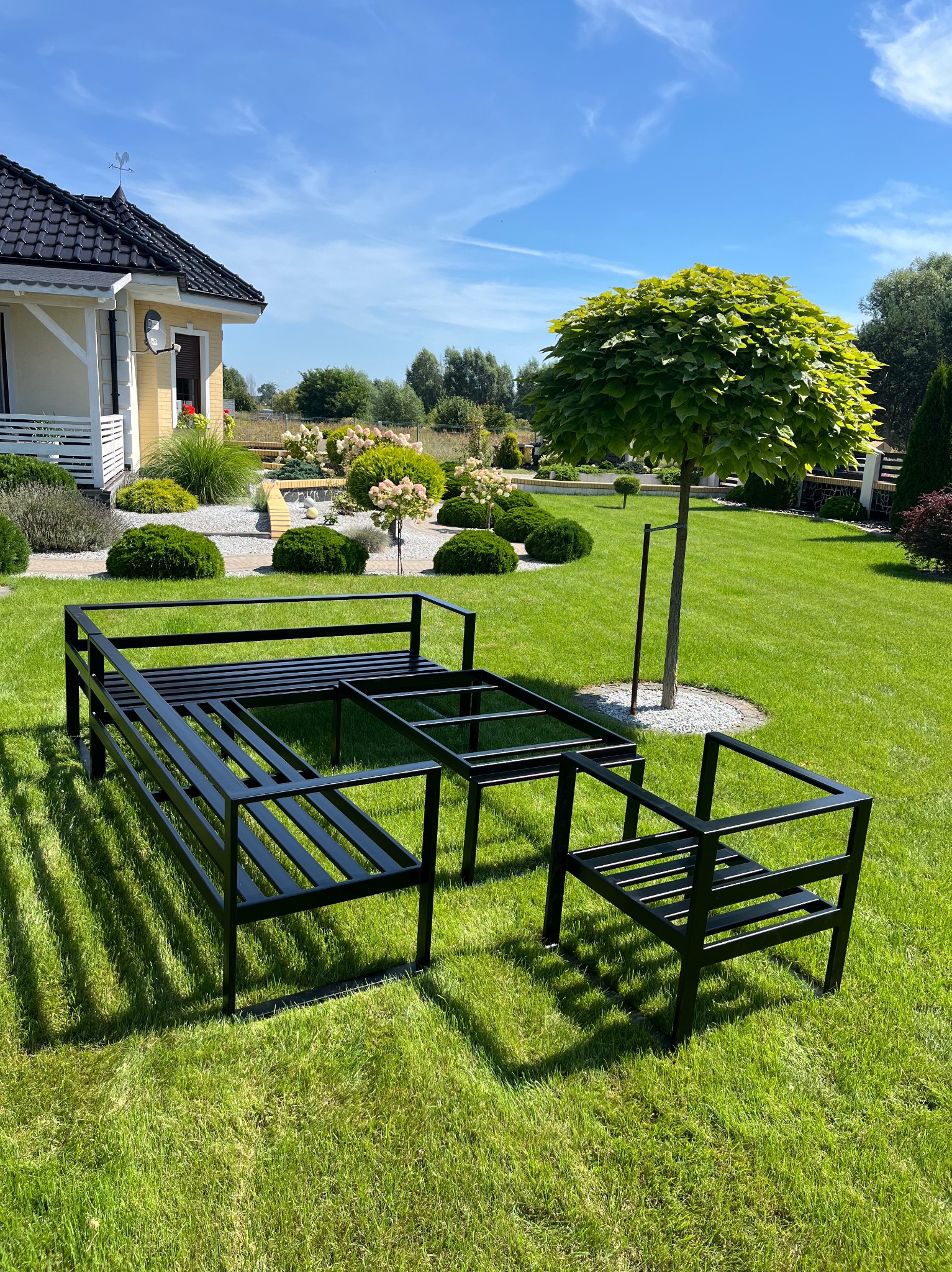 Stelaż metalowy mebli ogrodowych w kolorze czarnym, składający się z sofy, stołu i krzesła, ustawiony na trawniku przed domem z zadbanym ogrodem w słoneczny dzień.